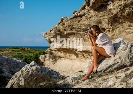 Vue latérale d'une femme cintrée bronzée en chemise blanche prenant des photos confortablement placée sur un rocher pierreux par temps lumineux Banque D'Images