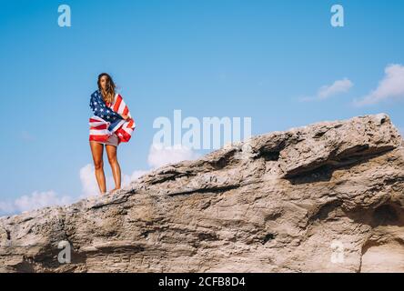 Gaie femme bronzée en vacances dans une ambiance décontractée tandis qu'une chemise se tient sur une grande falaise et tient le drapeau américain sous la main avec le ciel bleu et l'obélisque sur le fond Banque D'Images