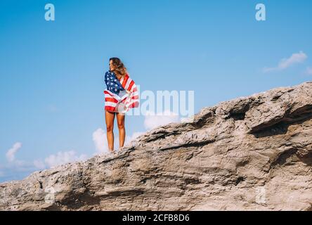 Gaie femme bronzée en vacances dans une ambiance décontractée tandis qu'une chemise se tient sur une grande falaise et tient le drapeau américain sous la main avec le ciel bleu et l'obélisque sur le fond Banque D'Images