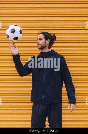 Jeune homme barbu de football en bleu foncé à capuche de sport et short tournant la balle sur le doigt tout en se tenant contre le rouleau jaune arrière-plan de la navette Banque D'Images