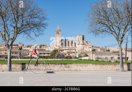 De dessous vue latérale de mâle méconnaissable dans des vêtements décontractés monter à vélo rétro sur la grande roue le long de la rue contre les façades de petits bâtiments et ancien palais à l'horizon dans le centre-ville sous ciel bleu sans nuages par temps ensoleillé Banque D'Images