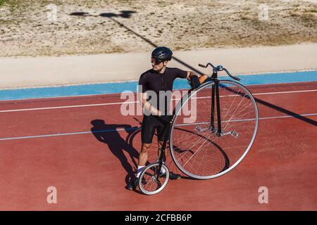 D'en haut sérieux homme en casque d'activité noir loin en vous tenant debout avec le vélo rétro penny farthing sur piste de course au terrain de sport Banque D'Images