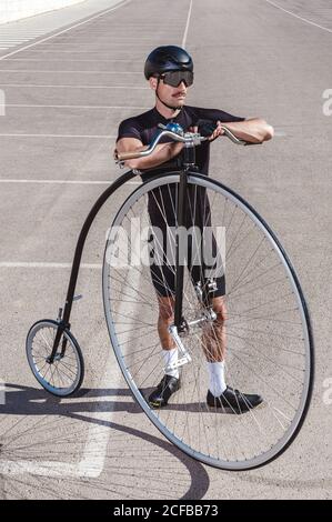 De dessus l'homme pensif en surcombinaison noire et le casque regardant loin en se tenant dans le parking avec la roue rétro haute vélo contre mur en béton par temps ensoleillé Banque D'Images