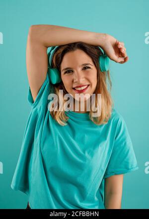Bonne jeune femme en t-shirt lumineux souriant regardant l'appareil photo et écoutant de la musique dans les écouteurs sur fond turquoise Banque D'Images
