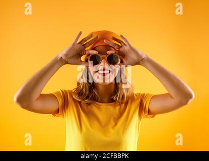 Bonne jeune femme en vêtements brillants et lunettes de soleil stylées souriant et regardant l'appareil photo sur fond orange Banque D'Images