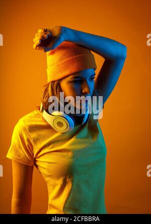 Jeune femme pensive en bonnet orange avec casque autour du cou en maintenant le bras au-dessus de la tête et en regardant loin sur fond orange Banque D'Images