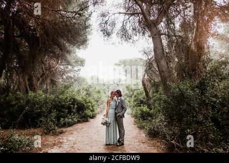 Jeune couple récemment marié dans le port de mariage debout et embrassant sur le chemin entre belle forêt verte avec de grands arbres Banque D'Images