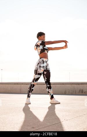 D'en-dessous, une femme adulte afro-américaine dans des vêtements de sport fleuris qui étirent les muscles des mains tout en restant seule et en se réchauffant avant de s'entraîner dans un environnement urbain par beau temps Banque D'Images