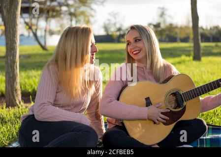 Deux filles blondes se réjouissent ensemble, chantant et jouant de la guitare dans l'herbe. Pique-nique musical dans le parc pendant une journée ensoleillée. Banque D'Images