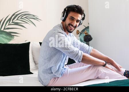 Vue latérale d'un homme hispanique barbu dans un casque assis sur le lit et profiter de la bonne musique à la maison appareil photo Banque D'Images