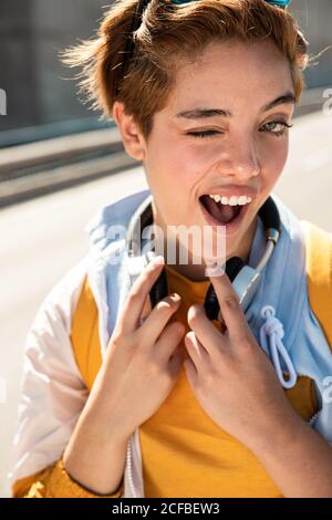 Magnifique femme millénaire élégante et élégante, avec ses yeux verts étonnants et son sourire éclatant dans des vêtements blancs et jaunes avec casque sans fil et lunettes de soleil, regardant l'appareil photo contre une route floue par beau temps Banque D'Images
