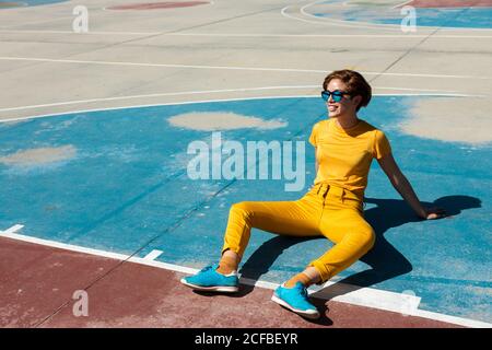 Grand angle de frowning froide femme adolescente dans des vêtements jaunes avec des lunettes de soleil tout en étant assis seul sur un terrain de sport bleu de basket-ball contre les bâtiments modernes flous à plusieurs étages en été Banque D'Images