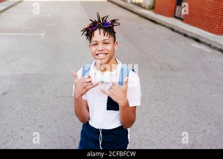 Jeune homme afro-américain joyeux et élégant en vêtement décontracté avec casque debout dans la rue et montrant le panneau de la main shaka Banque D'Images