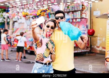 Joyeuse femme dans des lunettes de soleil et des vêtements décontractés à l'aide d'un smartphone de prise selfie avec le gars tout en mangeant des bonbons pomme et coton sucreries sur la foire Banque D'Images