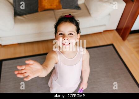 D'au-dessus de la petite fille avec ruban souriant pour l'appareil photo et main agitant pendant l'entraînement gymnastique rythmique à la maison Banque D'Images