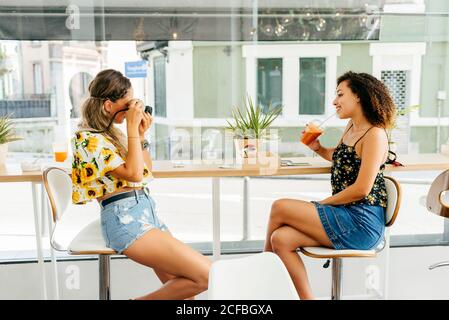 Vue latérale d'une jeune femme qui prend en photo une femme ethnique ami avec une boisson saine tout en se reposant dans un restaurant élégant ensemble