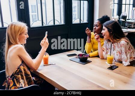 Vue latérale d'une jeune femme blonde heureuse prenant des photos avec le smartphone de diverses amies qui mangent du smoothie tout en s'asseyant ensemble à table dans le café et en prenant le petit déjeuner Banque D'Images