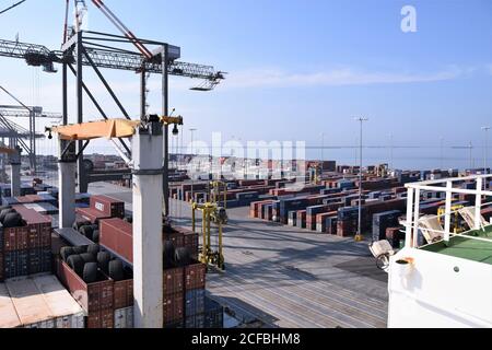 Vue depuis le pont du navire marchand sous opération de fret sur un quai de terminal à conteneurs recouvert de conteneurs prêts à être chargés sous ciel bleu. Banque D'Images