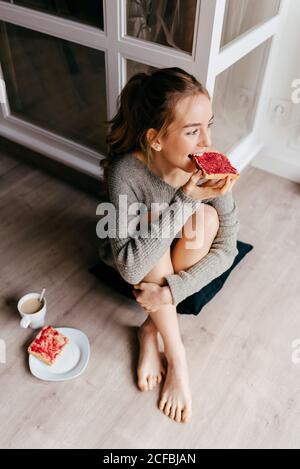 De dessus de positive jeune femme dans l'habillement décontracté ayant petit déjeuner avec pain grillé et une tasse de café tout en étant assis sur le sol dans la chambre Banque D'Images