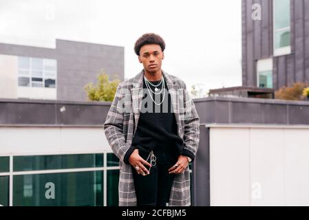 Un jeune homme à la mode afro-américain sérieux en manteau à carreaux tendance et un pantalon serré qui regarde l'appareil photo tout en se tenant contre un vêtement moderne bâtiments de la ville Banque D'Images