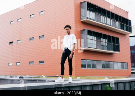 Vue d'en dessous de côté de jeune homme afro-américain élégant regarder l'appareil photo dans une chemise blanche et un pantalon serré avec baskets blanches sur la clôture contre le bâtiment rose de la ville Banque D'Images