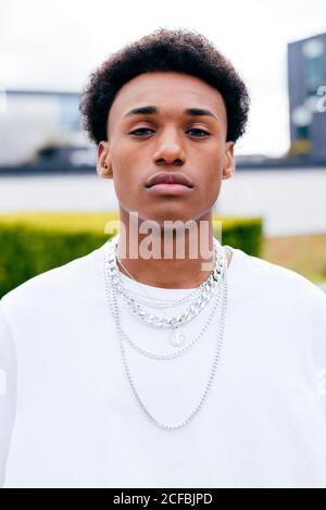 Jeune homme aux cheveux bouclés noirs adolescent dans des vêtements élégants avec chaînes regardant la caméra en se tenant sur la rue de la ville avec plantes vertes et bâtiments en arrière-plan Banque D'Images