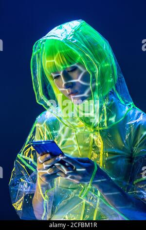 Jeune femme asiatique en tenue futuriste et perruque verte utilisant un smartphone en lumière fluorescente Banque D'Images