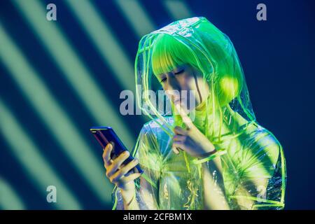 Jeune femme asiatique en tenue futuriste et perruque verte utilisant un smartphone en lumière fluorescente Banque D'Images