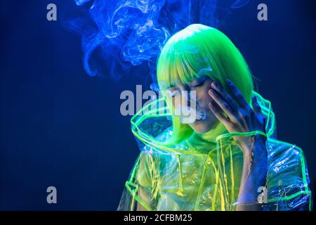 Jeune femme asiatique assez inhabituelle en plastique transparent imperméable et cheveux jaunes fumer dans la lumière fluorescente Banque D'Images