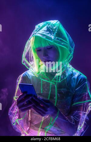 Jeune femme asiatique en tenue futuriste et perruque verte utilisant un smartphone en lumière fluorescente Banque D'Images