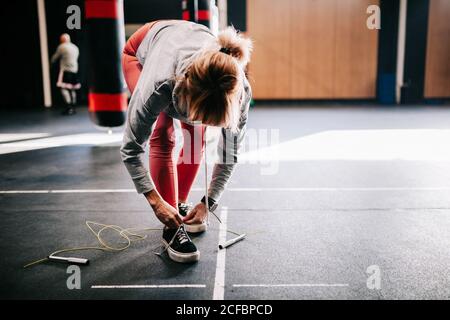 athlète féminine non reconnaissable dans les vêtements de sport qui nouent des lacets sur les baskets pendant se tenir près d'un sac de poinçonnage suspendu et se préparer à l'entraînement salle de sport moderne Banque D'Images