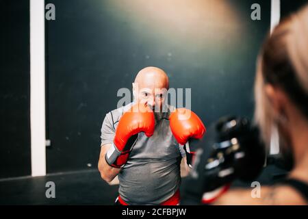 Vue arrière d'un jeune sportif écourté et méconnu en gants de boxe en train de s'entraîner en compagnie d'un entraîneur masculin professionnel dans une salle de sport moderne Banque D'Images