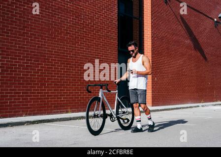 Cycliste masculin élégant en vêtements de sport et lunettes de soleil en utilisant le smartphone pendant debout avec un vélo près du mur de briques rouges Banque D'Images