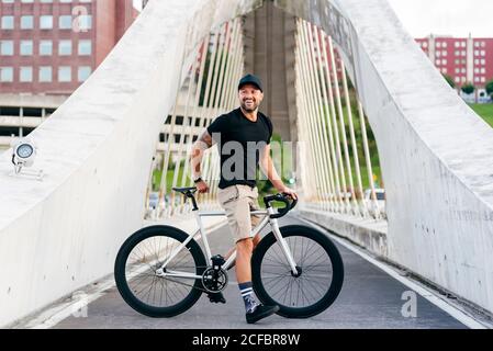 Joyeux homme barbu adulte en casquette noire portant une chemise noire et un short beige debout avec vélo à travers la passerelle dans la ville vue sur l'extérieur Banque D'Images