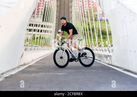 Homme barbu adulte en casquette noire portant une chemise noire et short beige à vélo traversant la passerelle à la recherche de la ville loin Banque D'Images