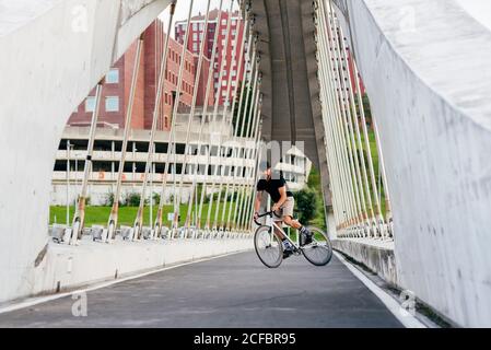 Homme barbu adulte en casquette noire portant une chemise noire et short beige à vélo traversant la passerelle à la recherche de la ville loin Banque D'Images