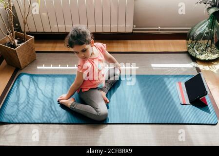 De dessus positive petite fille dans le yoga pose sur le tapis près de la fenêtre à la maison tout en pratiquant le yoga et en regardant la vidéo didacticiels sur tablette Banque D'Images