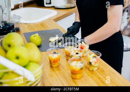 Crop Woman en tablier noir et gants hacher des pommes vertes fraîches avec un couteau et préparer des desserts frais sains dans la cuisine moderne Banque D'Images