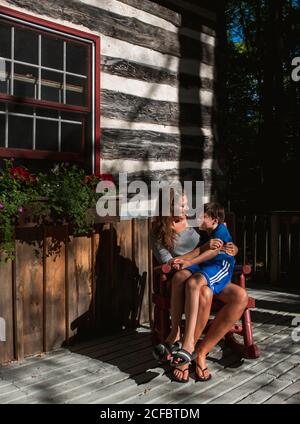 Mère et fils assis dans une chaise à bascule sur le pont d'une cabane rustique en bois. Banque D'Images