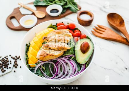 Du poulet frit et des tranches de mangue placées près de la cerise tomates et moitié d'avocat sur les feuilles d'épinards et l'oignon sur une table en marbre près des ustensiles et des sauces aux épices Banque D'Images