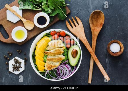 Vue de dessus du poulet frit et des tranches de mangue placées à proximité tomates cerises et moitié d'avocat sur les feuilles d'épinards et oignon sur table en marbre gris près des ustensiles et sauces avec épices Banque D'Images