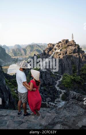 De l'arrière vue de couple de touristes dans traditionnel chapeaux coniques debout sur une colline rocheuse et bénéficiant d'une vue majestueuse De la grotte de Mua tout en se câlinant et en regardant l'un l'autre Banque D'Images