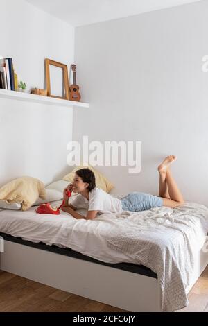 Jeune femme heureuse souriant et répondant à un appel téléphonique de vintage téléphone rouge tout en étant allongé sur le lit dans la chambre confortable Banque D'Images