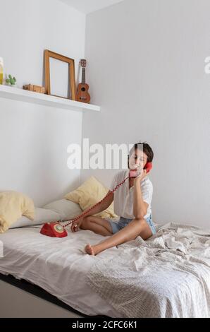 Jeune femme heureuse souriant et répondant à un appel téléphonique de vintage téléphone rouge tout en étant assis sur le lit dans la chambre confortable Banque D'Images