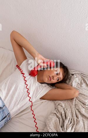 Jeune femme attentionnés répondant à un appel téléphonique depuis un téléphone rouge vintage tout en étant allongé sur le lit dans une chambre confortable Banque D'Images