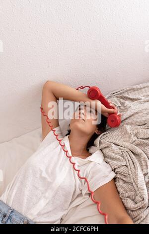 Par le dessus, une femme enfoncée avec un combiné téléphonique rouge près de la tête regarder pendant une conversation téléphonique sur le lit Banque D'Images