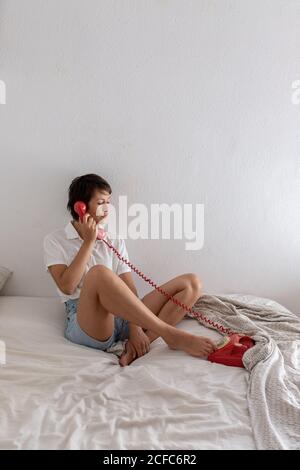 Jeune femme et répondre à un appel téléphonique depuis un téléphone rouge vintage tout en étant assis sur un lit dans une chambre confortable Banque D'Images