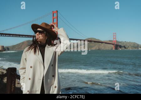 Vue latérale d'une jeune femme souriante dans une tenue tendance avec chapeau et lunettes de soleil debout sur une rive contre le Golden Gate Bridge en Californie par beau temps Banque D'Images