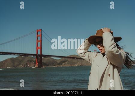 Vue latérale de la jeune femme en tenue tendance avec chapeau et lunettes de soleil debout sur le remblai contre le Golden Gate Bridge en Californie par temps ensoleillé Banque D'Images