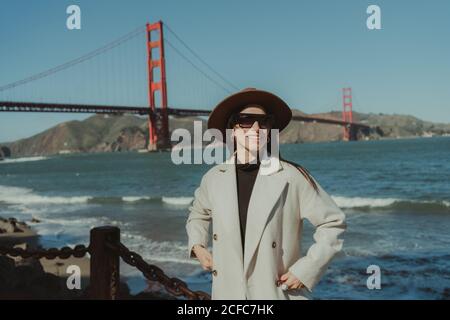 Vue latérale d'une jeune femme souriante dans une tenue tendance avec chapeau et lunettes de soleil debout sur une rive contre le Golden Gate Bridge en Californie par beau temps Banque D'Images